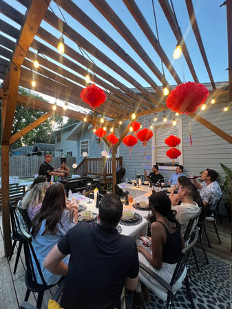 Twilight backyard dinner party on a wooden pergola-covered deck with hanging string lights and red paper lanterns, guests seated around a long table at a suburban home.