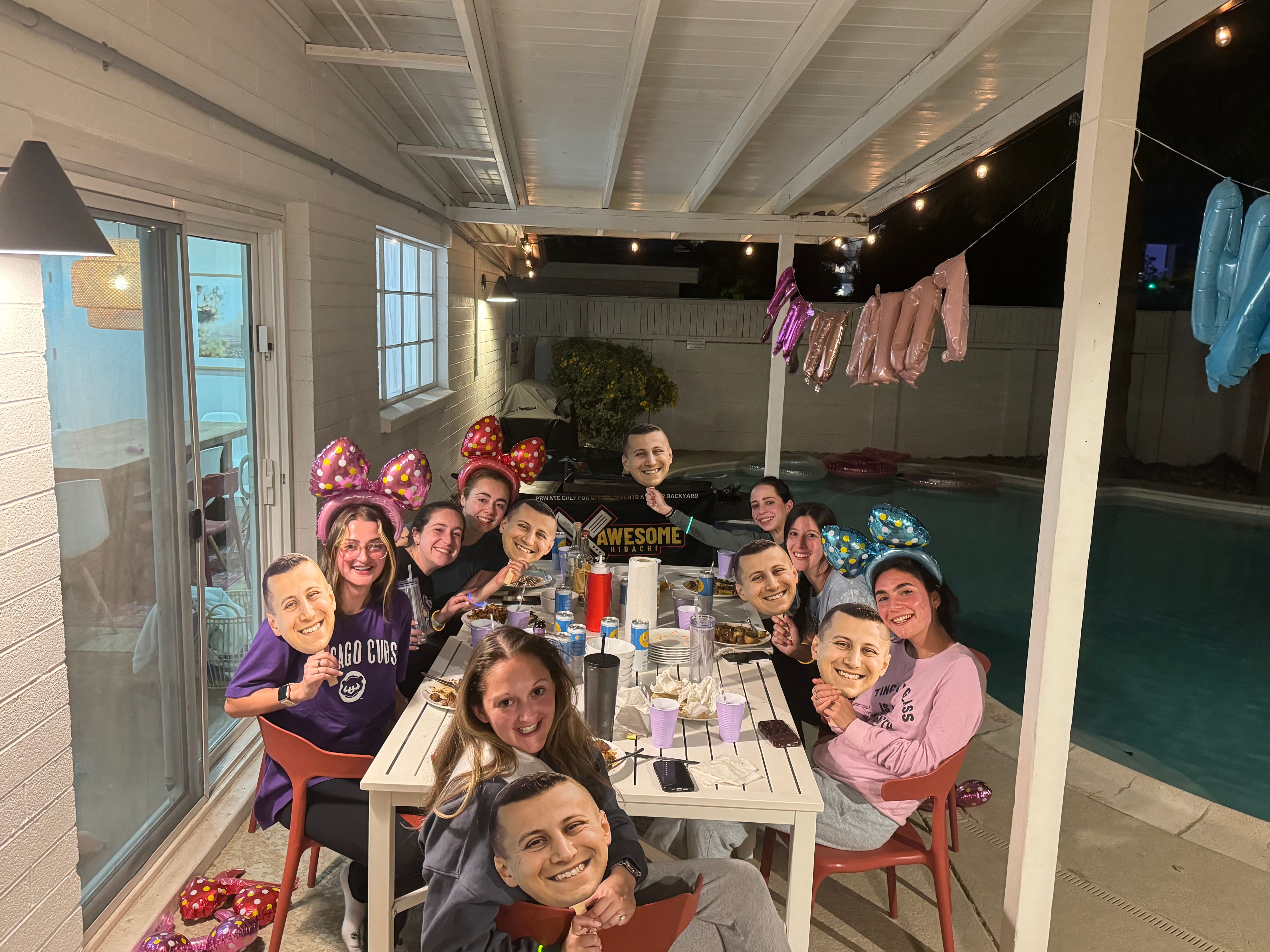Group of friends at a nighttime backyard poolside patio dinner under a covered pergola, seated around a long table with plates and drinks, wearing colorful bow headbands and holding oversized smiling face cutout masks, with string lights, metallic balloons and pool floats visible.