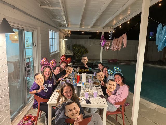 Group of friends at a nighttime backyard poolside patio dinner under a covered pergola, seated around a long table with plates and drinks, wearing colorful bow headbands and holding oversized smiling face cutout masks, with string lights, metallic balloons and pool floats visible.