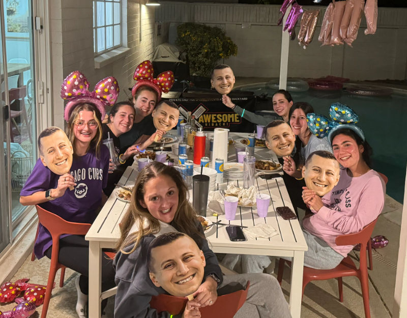 Cheerful group of friends at a backyard patio pool party around a long outdoor dining table at night, many wearing colorful polka-dot bow headbands and holding oversized smiling face cutout masks, with plates, cups and party balloons visible.