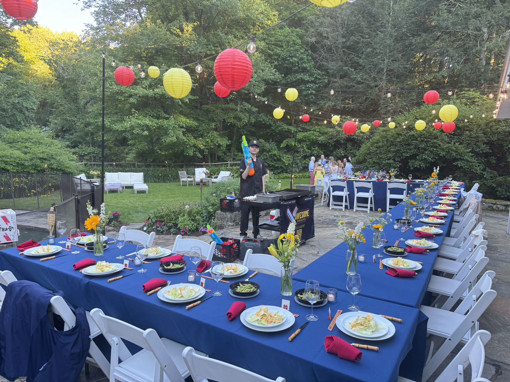 Backyard garden party on a stone patio — long blue-tablecloth banquet tables with place settings, white folding chairs, red and yellow paper lanterns and string lights.