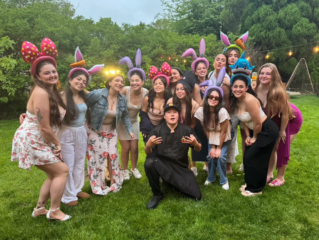 Smiling group of friends on a green backyard lawn at an outdoor evening party, many wearing colorful bunny-ear and novelty headbands with string lights and trees behind them.