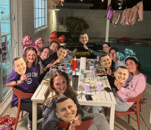 Group of friends at a nighttime backyard poolside dinner party, seated around a white table with plates and drinks, wearing colorful novelty mouse-ear headbands and holding oversized cutout face masks, with string lights and pink balloons overhead.