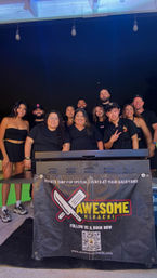 Smiling group of people and chefs posing behind an outdoor hibachi grill at a nighttime backyard party with hanging string lights and a promotional banner on the grill.