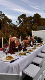 Outdoor backyard dinner table at dusk with white tablecloth, fall floral centerpieces, twisted taper candles, wine bottles, plated salads and white folding chairs against a treed sky
