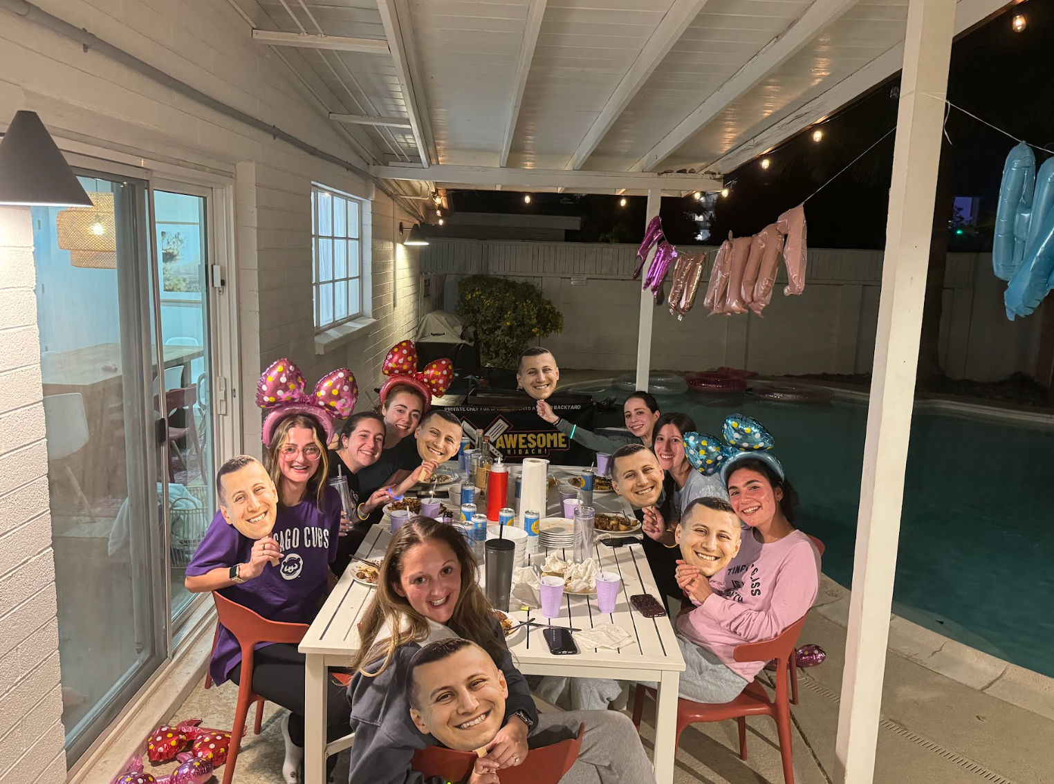 Backyard poolside birthday party on a covered patio at night — friends gathered around a table with food, balloons and string lights, wearing colorful bow headbands and holding identical face masks.