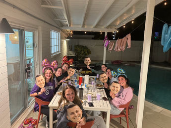 Backyard poolside birthday party on a covered patio at night — friends gathered around a table with food, balloons and string lights, wearing colorful bow headbands and holding identical face masks.