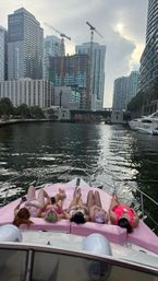Five people in colorful swimsuits lounging on pink cushions at the bow of a yacht on a Miami downtown waterway, with high‑rise skyline, construction cranes and an overcast sky.