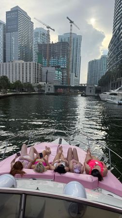 Five people in colorful swimsuits lounging on pink cushions at the bow of a yacht on a Miami downtown waterway, with high‑rise skyline, construction cranes and an overcast sky.