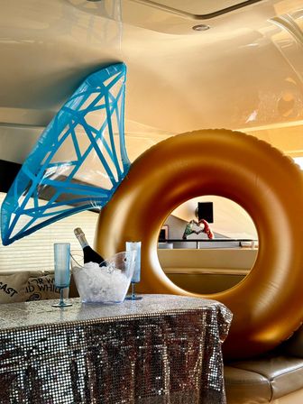 Luxurious yacht interior party setup with a large gold inflatable donut and blue mermaid-tail float, sequin-covered table holding an ice bucket with champagne and two blue flutes.