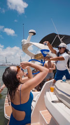 Yacht party scene: woman in a blue swimsuit drinks from a blue party funnel while a crew member pours, sunny day with harbor cranes and city skyline in the background.
