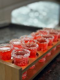 Close-up of bright red sugar-rimmed gelatin shots in clear cups arranged in a wooden shot flight tray on a dark countertop — festive party drinks.