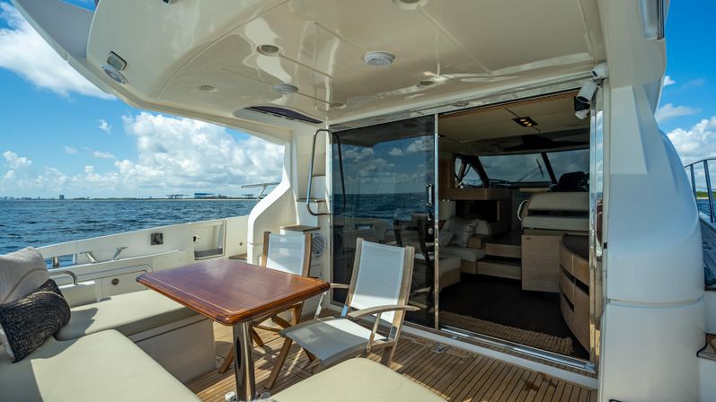 Luxury yacht aft deck with teak flooring, cushioned bench seating, wooden dining table and folding chairs, glass sliding doors to an interior salon and open sea view under a blue sky.