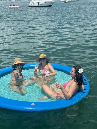 Three women in bikinis and sun hats relaxing and toasting with champagne glasses in a blue inflatable floating pool on a sunny bay with boats and yachts in the background