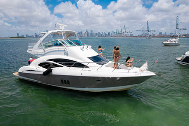 White luxury motor yacht with friends sunbathing on the bow in green coastal water, city skyline and cargo cranes on the horizon under a sunny blue sky