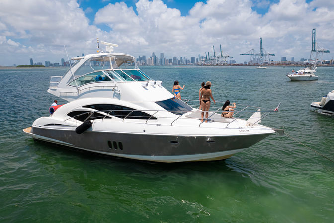 White luxury motor yacht with friends sunbathing on the bow in green coastal water, city skyline and cargo cranes on the horizon under a sunny blue sky