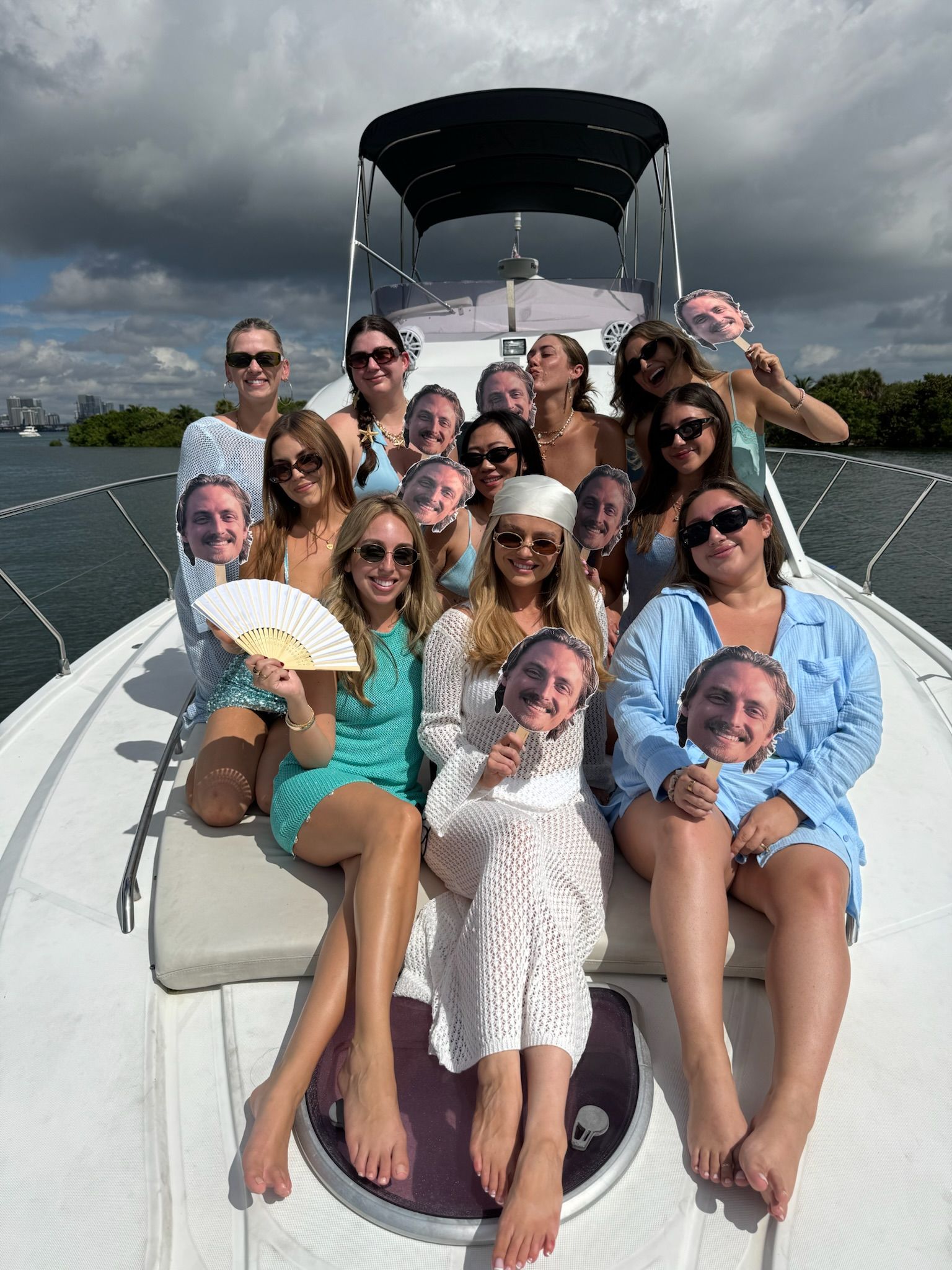 Yacht party: group of women on a boat in a coastal bay under dramatic clouds, posing and smiling while holding playful cutout face props