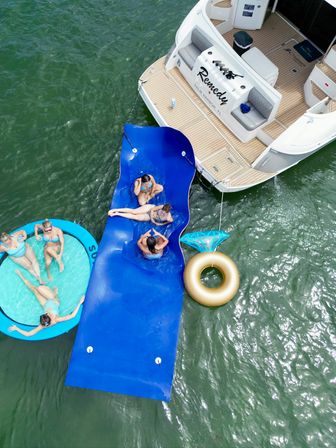 Aerial view of a white motorboat with a swim platform, an attached blue floating mat and round inflatable lounge where people in swimsuits relax on green water, plus a gold inner tube nearby.