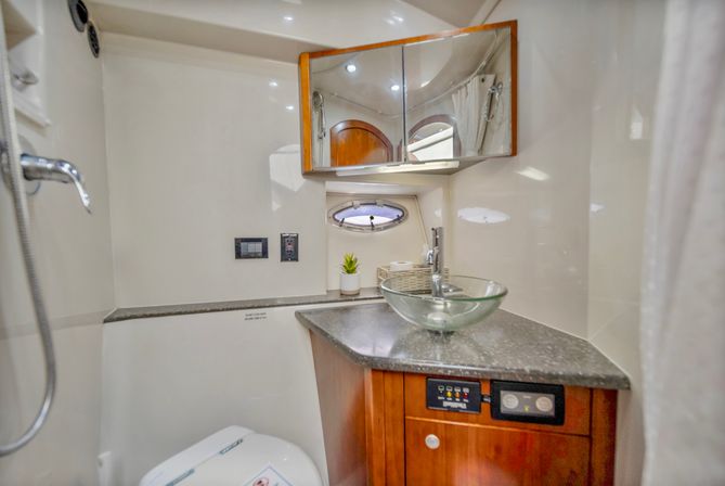 Compact yacht head with glass vessel sink on dark granite countertop, wooden vanity with control panel, mirrored cabinet, small porthole, handheld shower and marine toilet.
