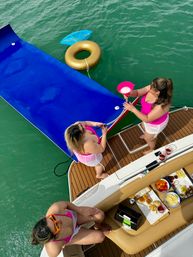 Summer boat party on emerald-green water: three people in pink swimwear on a motorboat swim platform with snacks and drinks, a blue floating slide and a gold donut inflatable nearby.