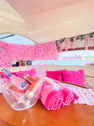 Bright pink-themed yacht interior with a floral wall, neon heart and “Let’s Go Girls” sign, hot-pink pillows and rolled towels, ice bucket of pink drinks on a wooden table, marina and water visible through the window.