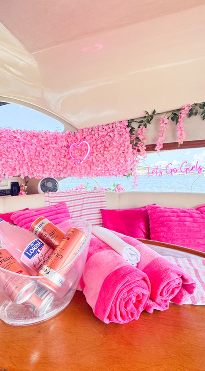 Bright pink-themed yacht interior with a floral wall, neon heart and “Let’s Go Girls” sign, hot-pink pillows and rolled towels, ice bucket of pink drinks on a wooden table, marina and water visible through the window.