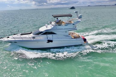 Luxury white motor yacht cruising turquoise coastal waters under a partly cloudy sky, leaving a foamy wake with passengers on the upper deck