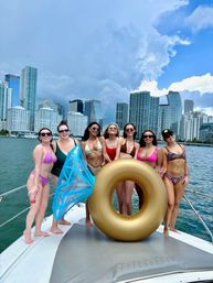 Seven people in colorful swimsuits posing on a yacht with a gold donut float, Miami skyline and dramatic clouds overhead