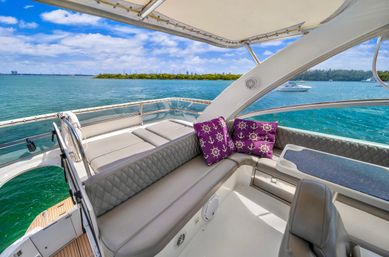 Sunlit luxury yacht deck with cushioned L-shaped seating, purple anchor-and-wheel pillows, and a small table overlooking turquoise bay, nearby mangrove island and distant coastal skyline under a blue sky.