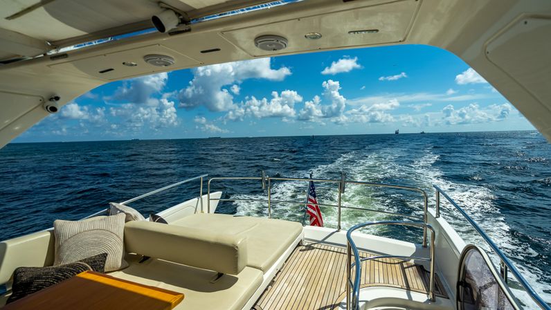 Sunlit yacht stern with teak deck and cushioned lounger, American flag fluttering as the boat leaves a foamy wake across the deep-blue open ocean under a bright sky with puffy clouds.