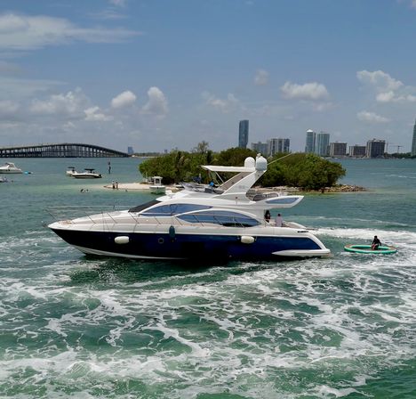 Luxury yacht cruising turquoise bay near a mangrove island, coastal city skyline and bridge under a sunny sky, with an inflatable raft trailing behind.