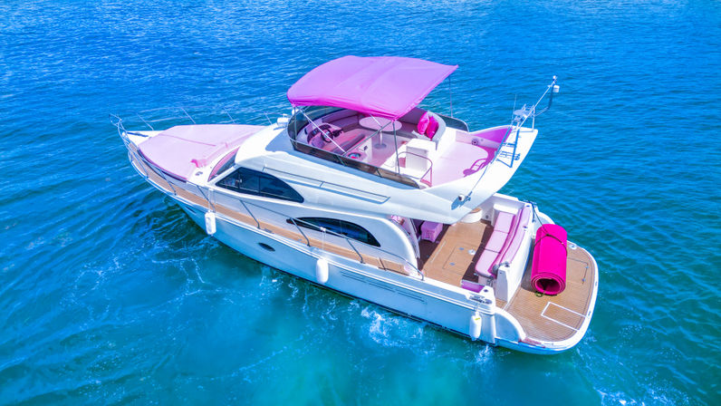 Aerial view of a white luxury motor yacht with bright pink cushions and canopy cruising on clear turquoise ocean water.