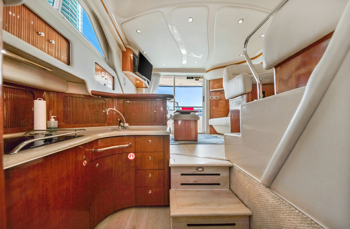 Interior of a luxury yacht galley and salon — glossy teak cabinets, compact sink, steps up to leather seating and marina view.