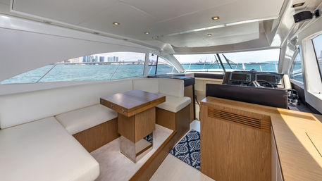 Sunlit luxury yacht interior salon with white leather seating, wooden foldable table and helm station, panoramic turquoise water and distant city skyline visible through wide windows