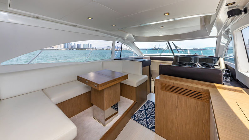 Sunlit luxury yacht interior salon with white leather seating, wooden foldable table and helm station, panoramic turquoise water and distant city skyline visible through wide windows