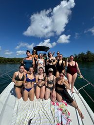 Group of women in swimsuits toasting on the bow of a motorboat during a sunny coastal boat party, blue sky with fluffy clouds and green shoreline in the background.