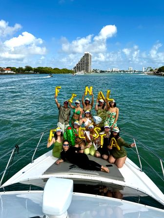Group of friends partying on a white yacht in turquoise coastal waters, holding gold "FINAL FIESTA" balloons with a waterfront city skyline and blue sky in the background.