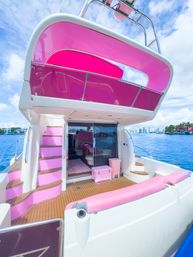 Bright pink-themed luxury motor yacht stern with teak deck, pink cushions and canopy, curved steps to an upper deck, clear blue water and a sunny tropical marina skyline in the distance.