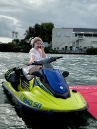 Playful woman in a white cover-up adjusting sunglasses while sitting on a yellow-and-blue jet ski on coastal water near palm trees and waterfront homes under a cloudy sky