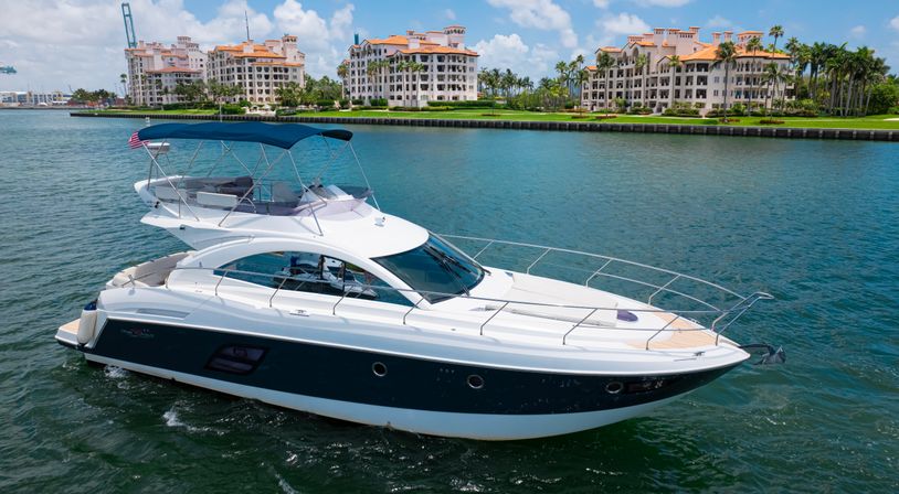 Sleek white luxury motor yacht cruising a turquoise inlet past Mediterranean-style waterfront condos and palm trees under a sunny blue sky