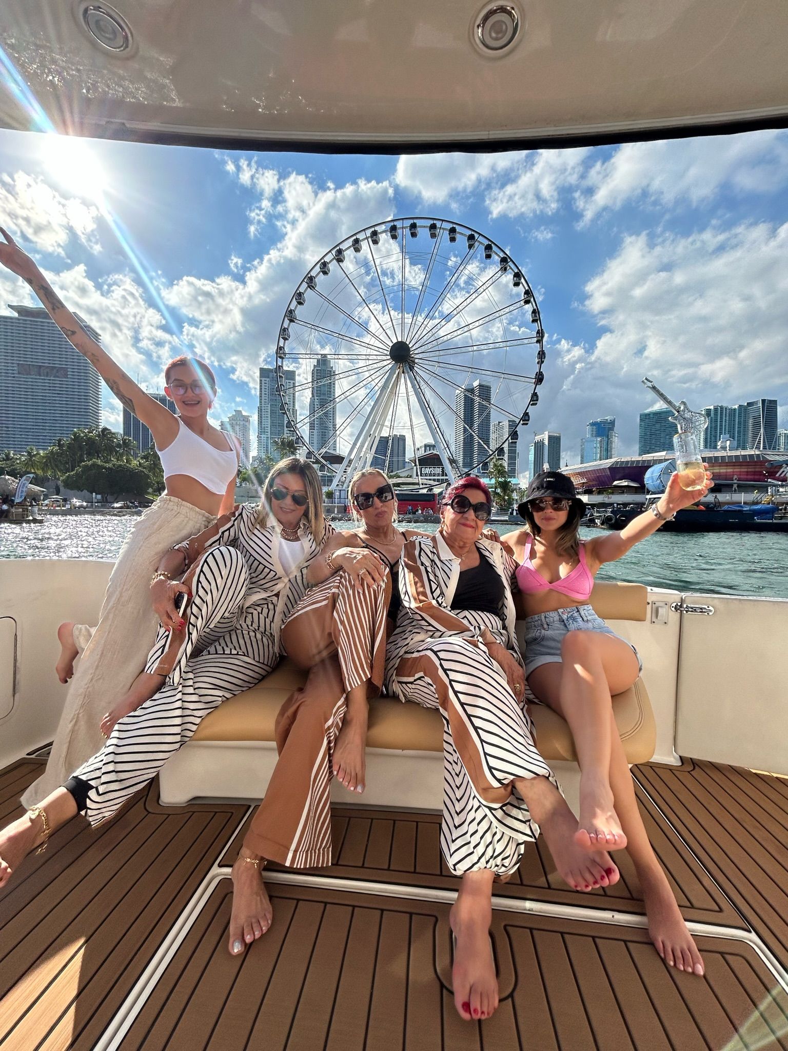 Five women relaxing barefoot on a yacht couch, posing with drinks against a waterfront city skyline and large Ferris wheel under a sunny blue sky