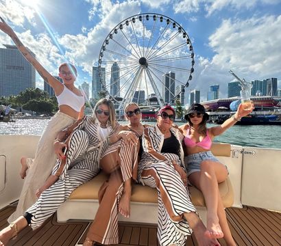 Five women relaxing barefoot on a yacht couch, posing with drinks against a waterfront city skyline and large Ferris wheel under a sunny blue sky