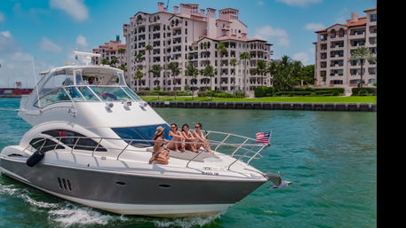 Friends sunbathing on the bow of a luxury motor yacht cruising turquoise coastal waters past palm-lined waterfront condos, American flag flying under a sunny sky.