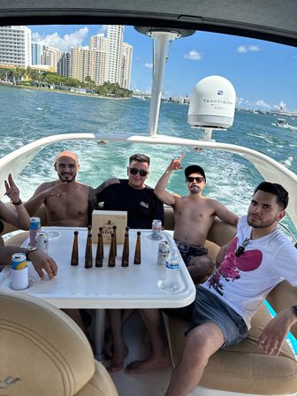Four friends enjoying a sunny yacht ride on turquoise bay with a high‑rise coastal skyline in the background, beers and small bottles on the table, relaxed party vibe.