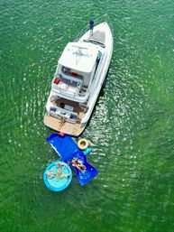 Aerial drone view of a white luxury yacht anchored in emerald-green water with sunbathers lounging on blue floating mats and a round inflatable pool beside the stern.