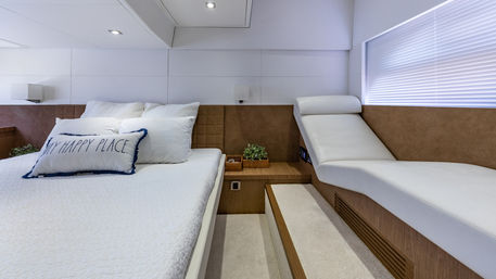 Cozy modern luxury yacht master cabin with white bedding, decorative “My Happy Place” pillow, wooden nightstand, chaise lounge and soft light through blinds