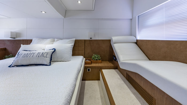 Cozy modern luxury yacht master cabin with white bedding, decorative “My Happy Place” pillow, wooden nightstand, chaise lounge and soft light through blinds