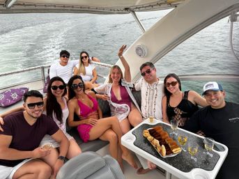 Cheerful group of friends relaxing on a motor yacht cruising open water, wearing swimsuits and sunglasses, gathered around a table with pastries and glasses of white wine, boat canopy overhead and wake trailing behind.
