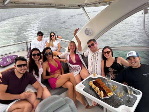 Cheerful group of friends relaxing on a motor yacht cruising open water, wearing swimsuits and sunglasses, gathered around a table with pastries and glasses of white wine, boat canopy overhead and wake trailing behind.