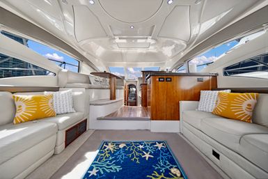 Luxury yacht salon interior with white leather sofas, yellow sunburst pillows, blue nautical rug with starfish and coral motifs, wood galley and helm ahead, and large windows framing marina and blue sky views